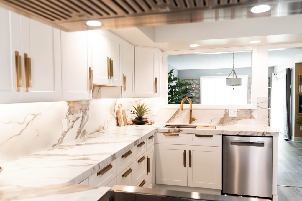Bright white kitchen with marble countertops, gold cabinet handles, and a stainless dishwasher under a marble island edge.