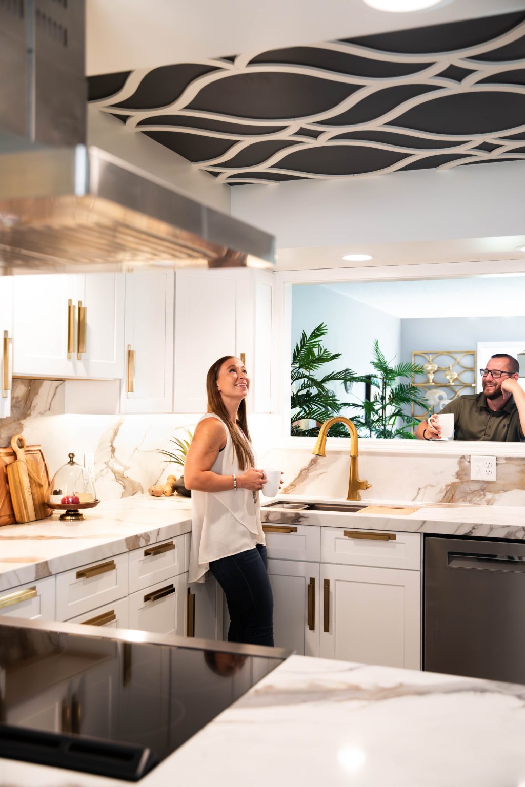 Two people chat in a bright modern kitchen by marble countertops, with plants visible in the background and gold-toned fixtures nearby.