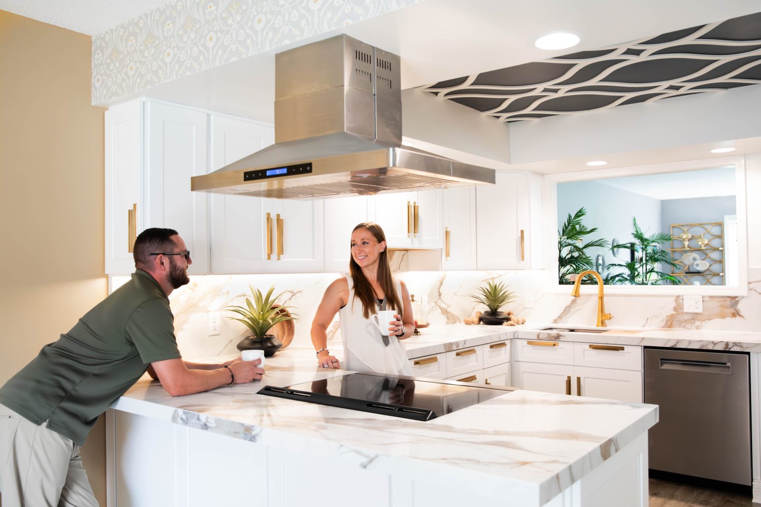 Two people chat at a white marble kitchen island with plants, white cabinets, and a stainless vent hood above the stove.