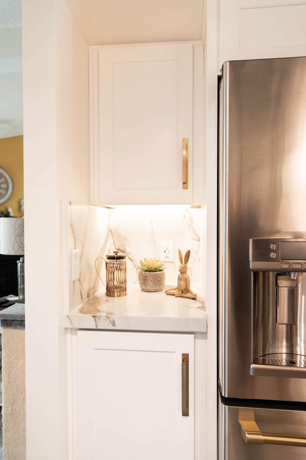 White upper and lower storage cabinets and marble countertop to the left of a stainless steel refrigerator