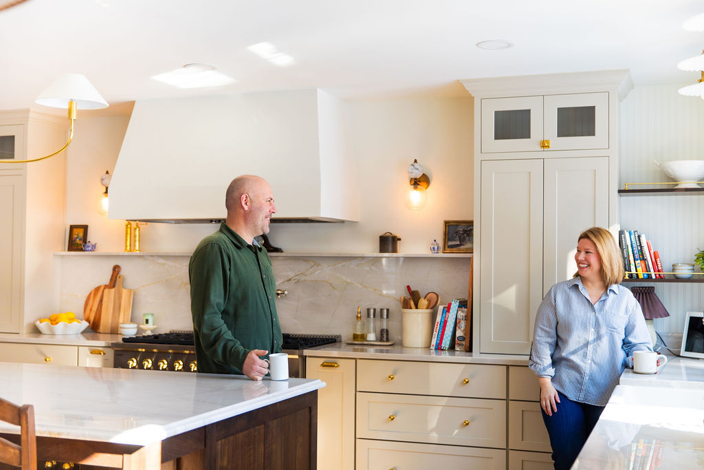 Two adults chatting in a bright cream kitchen, each holding a mug on a marble island.